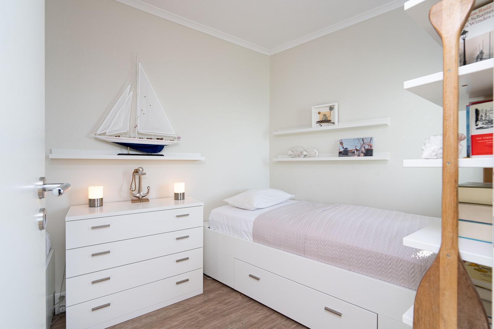 Bedroom with white bed and dresser, wall shelves with decor and model sailboat.