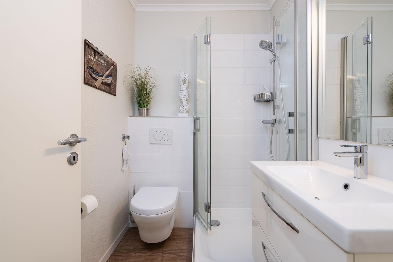 Bathroom with shower, sink, and toilet. White tiles and wooden floor.