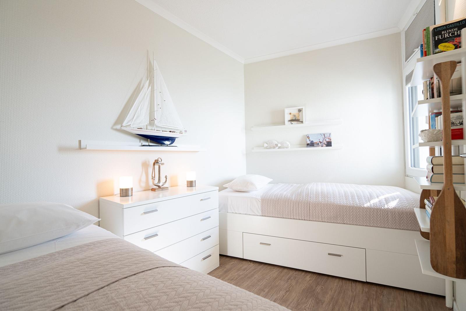 Room with two beds, dresser, and bookshelf with books and sailboat decoration.