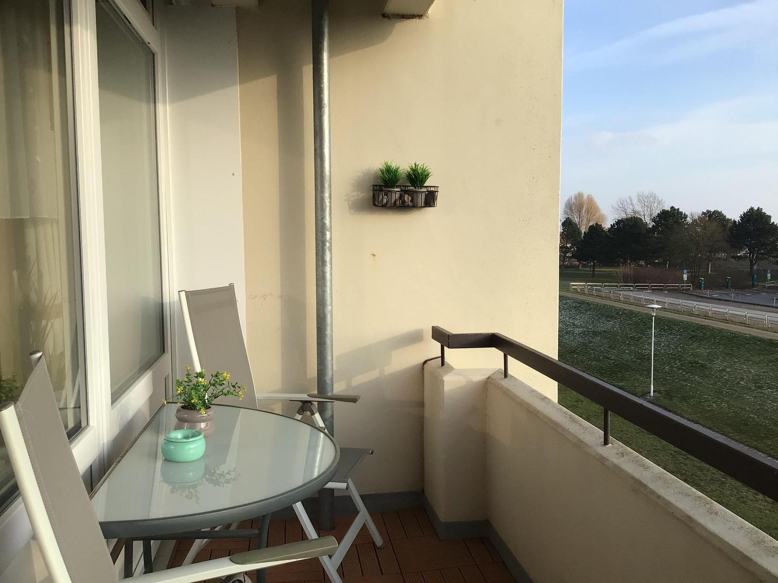 Balkon mit Tisch, Stühlen und Pflanzen. Blick auf Grünfläche und Straße.
