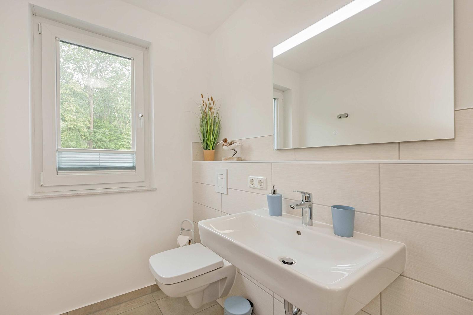 Badezimmer mit Waschbecken, Spiegel, Toilette und Fenster mit Blick auf Wald.