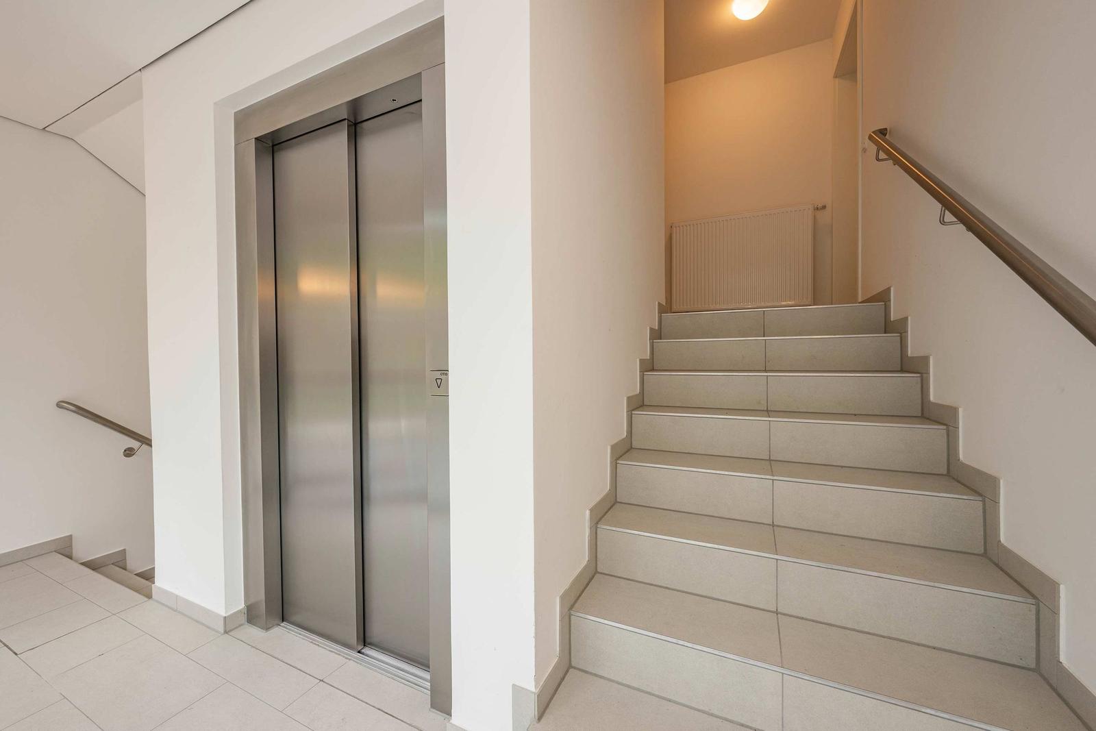 Elevator und Treppe im Eingangsbereich mit Metallgeländer und hellen Wänden.
