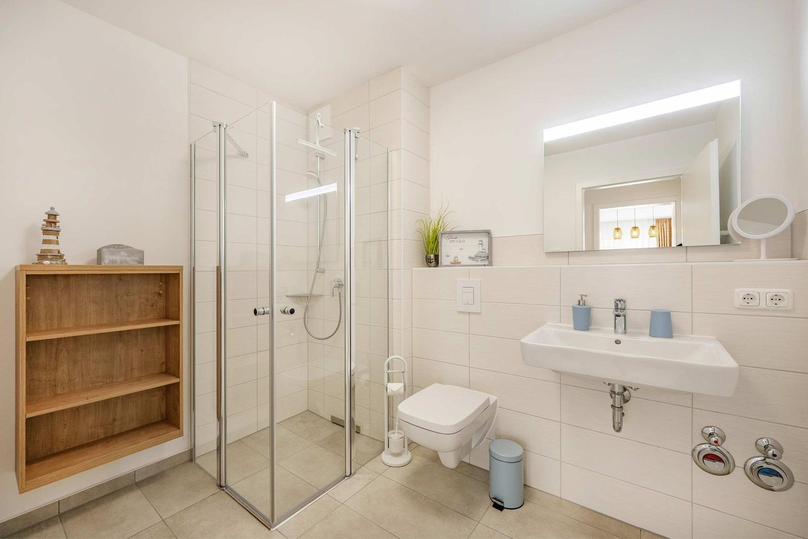 Badezimmer mit Dusche, Waschbecken und Toilette. Holzregal an der Wand.
