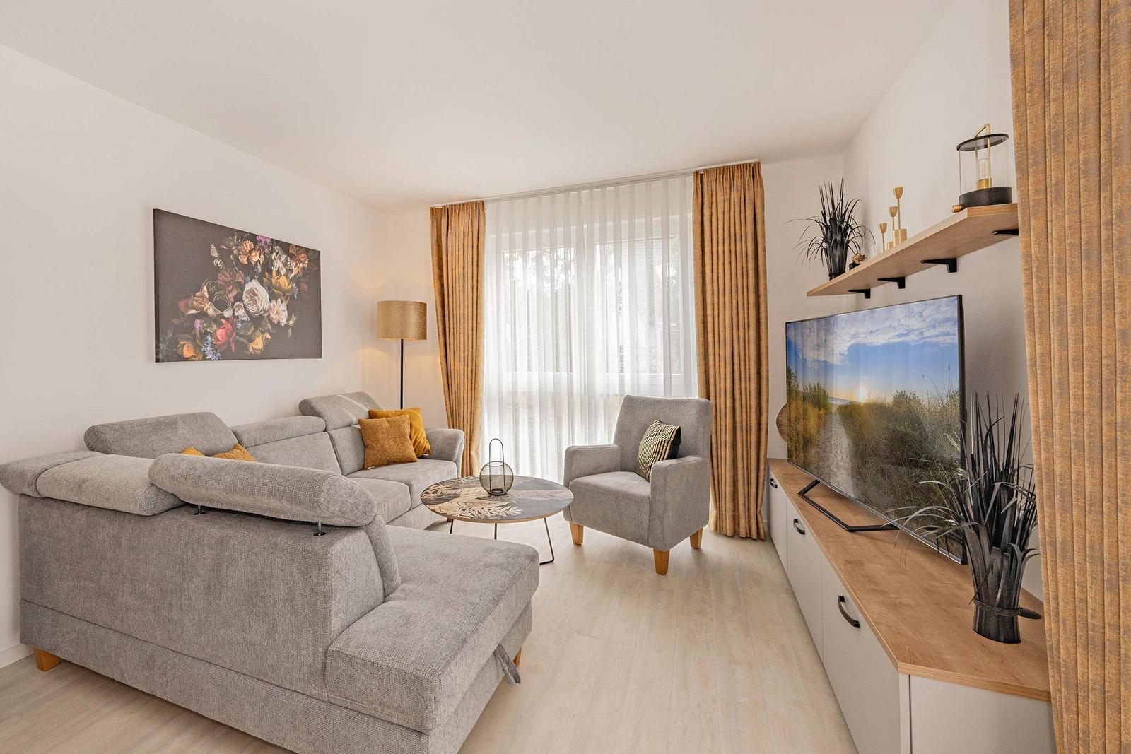 Wohnzimmer mit Sofa, Sessel, TV und Fenster mit Vorhängen.