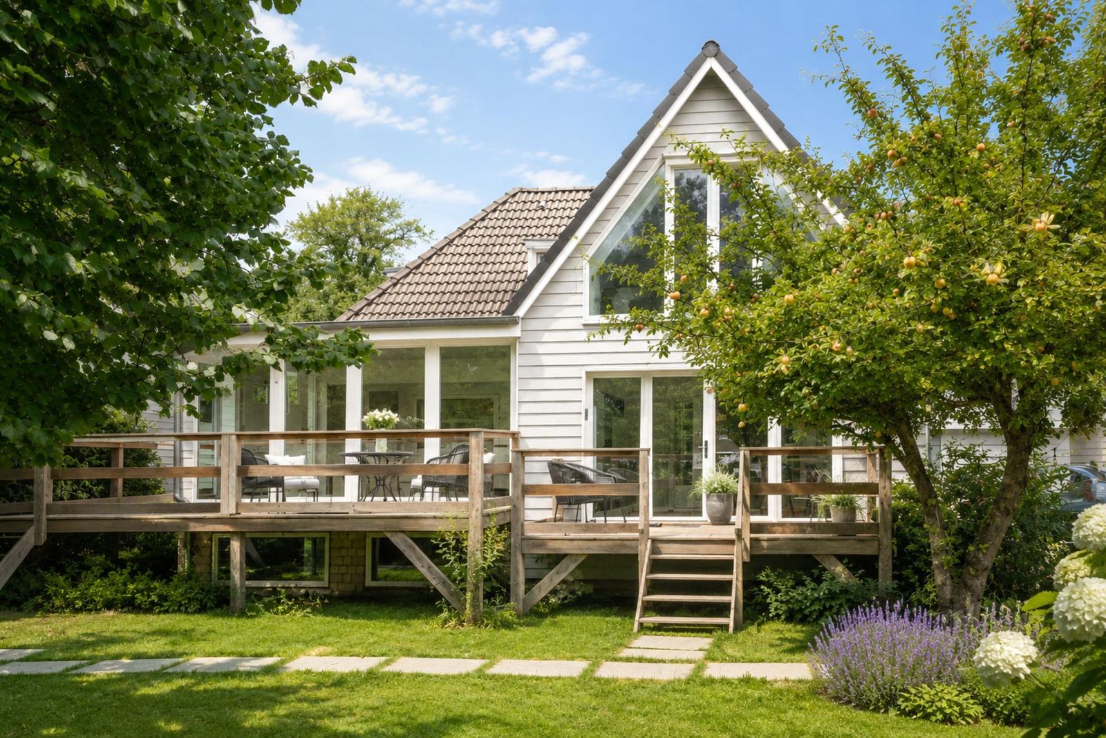 Ein weißes Haus mit Holzterrasse und Garten. Ein Apfelbaum steht vor dem Haus.
