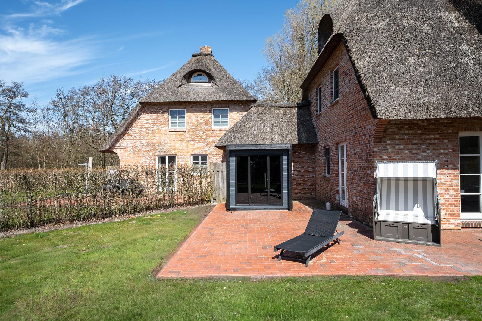 Rotes Backsteinhaus mit Strohdach, Terrasse mit Liegestuhl und Sonnenschutz.