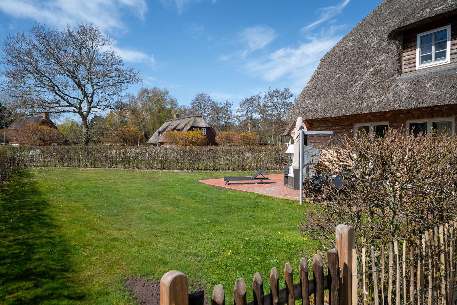 Haus mit Strohdach, Garten mit Liege und Terrasse unter blauem Himmel.
