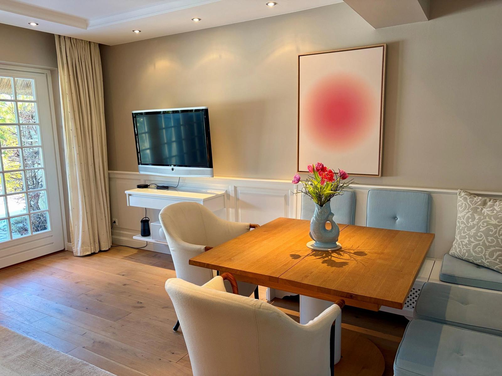 Esszimmer mit Holztisch, Stühlen und Fernseher an der Wand. Blumenvase auf Tisch. Fenster mit Vorhängen.