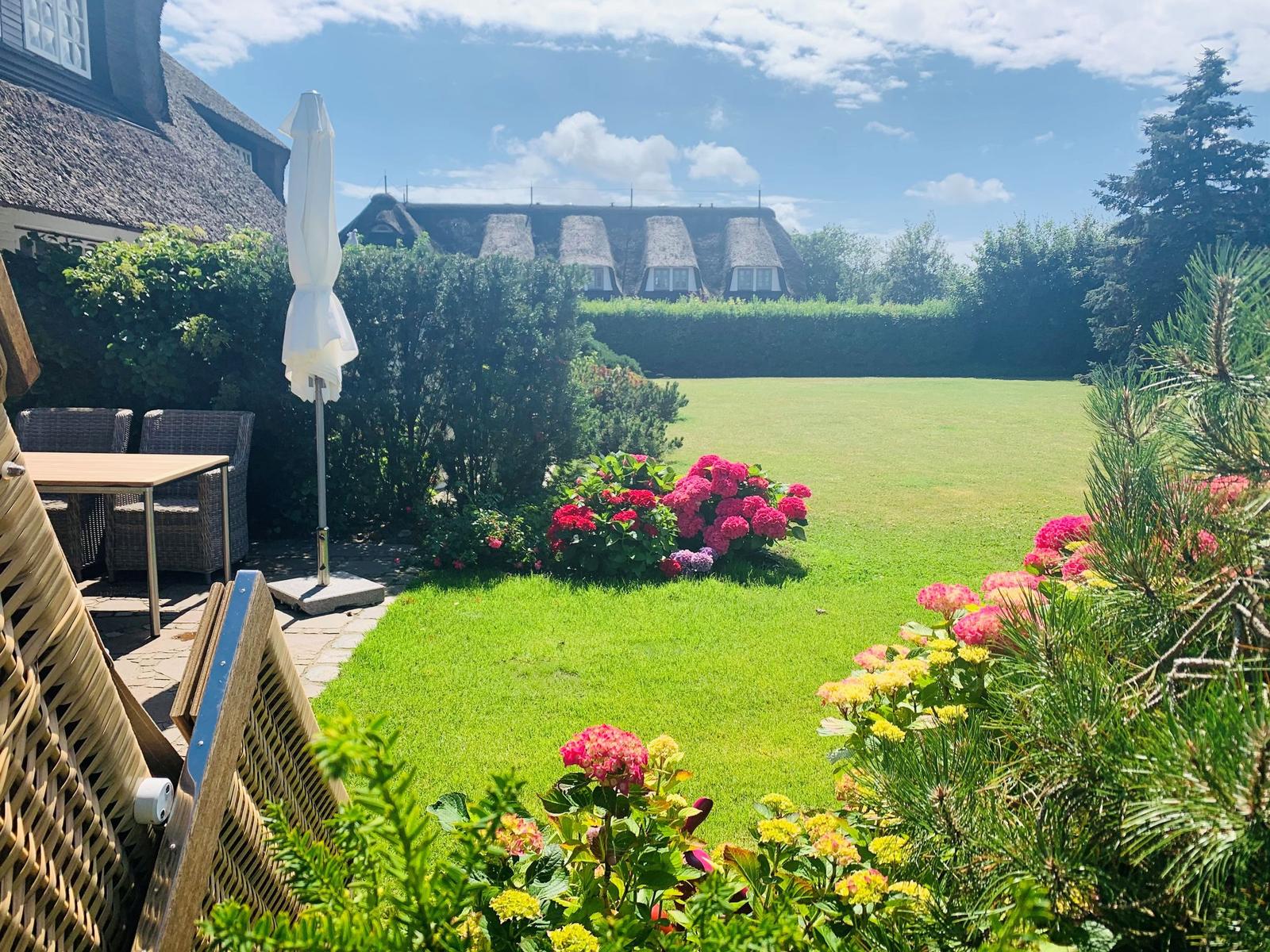 Garten mit Blumenbeeten, Rasenfläche und Terrasse mit Tisch und Stühlen.