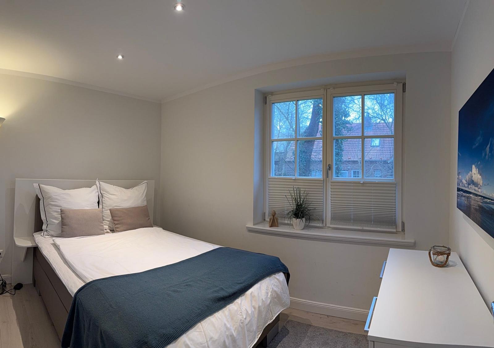 Schlafzimmer mit Bett, Fenster und Schreibtisch. Blick auf Gebäude und Bäume.