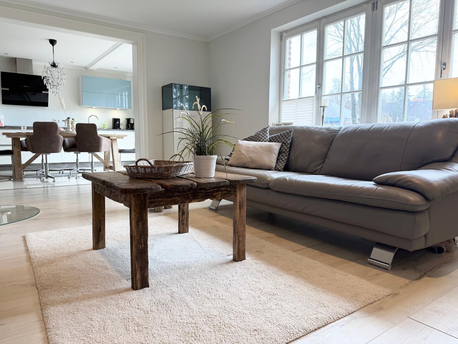 Ein Wohnzimmer mit grauem Sofa, rustikalem Tisch und großen Fenstern. Küche im Hintergrund.