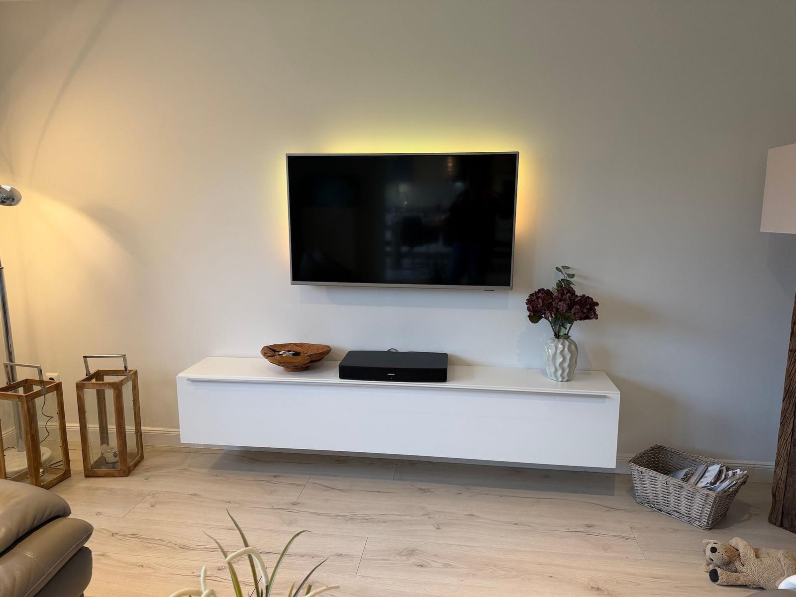 Wandmontierte TV mit Soundbar und Dekorationsgegenständen auf einem weißen Schwebetisch.