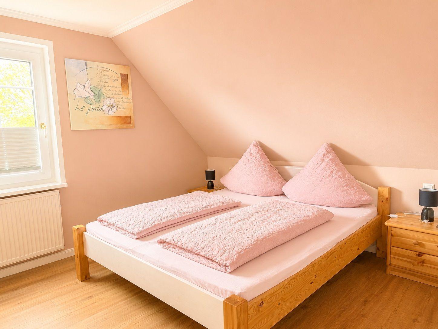 Schlafzimmer mit Doppelbett, Holzmöbel und Fenster. Wandfarbe rosa. Decke und Kissen rosa.