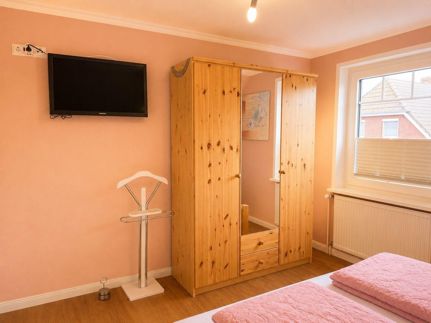 Schlafzimmer mit Bett, Kleiderständer, Schrank und Wandfernseher.