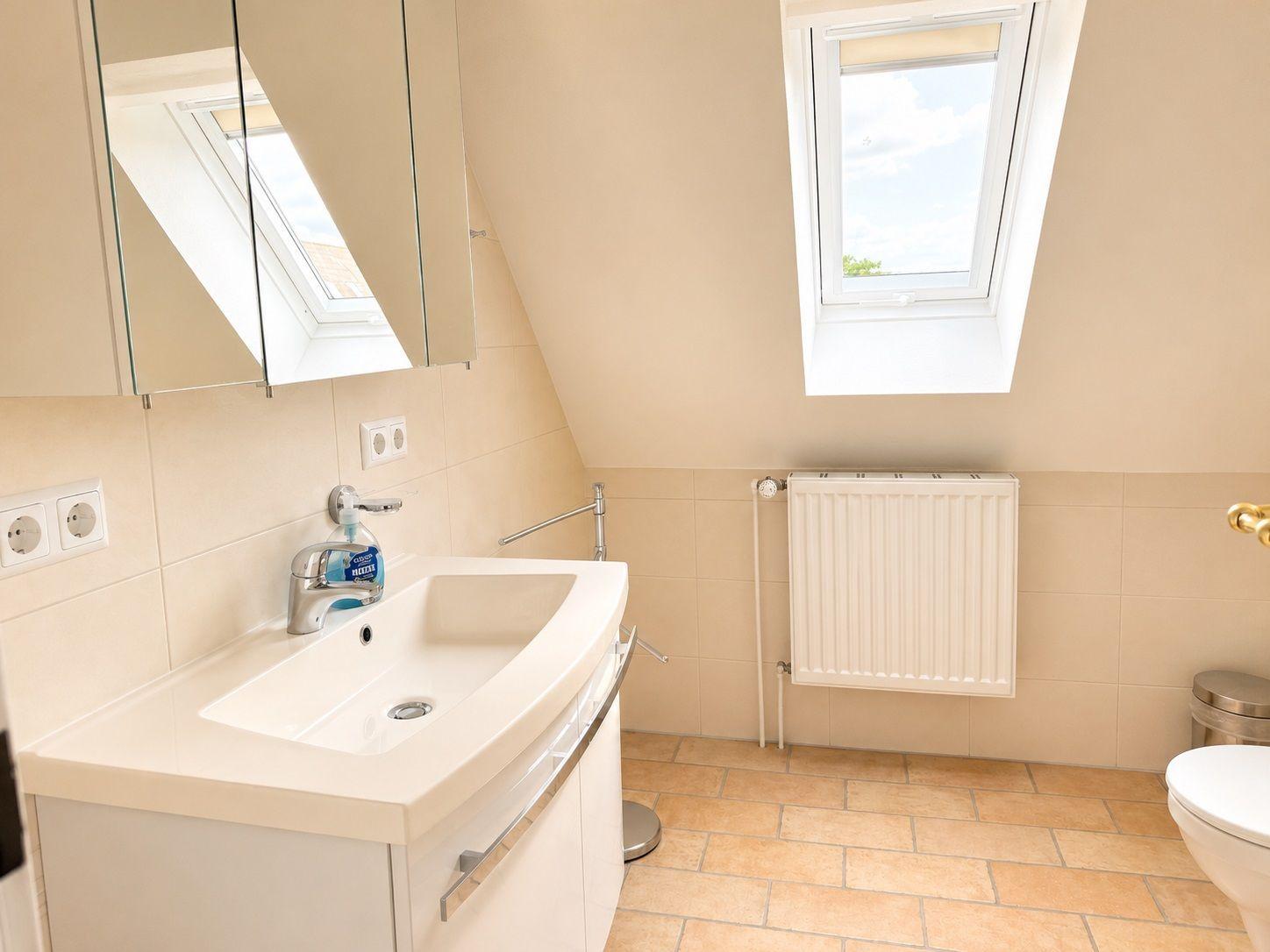 Badezimmer mit Waschbecken, Spiegel und Dachfenster. Bodenfliesen und Heizkörper sichtbar.