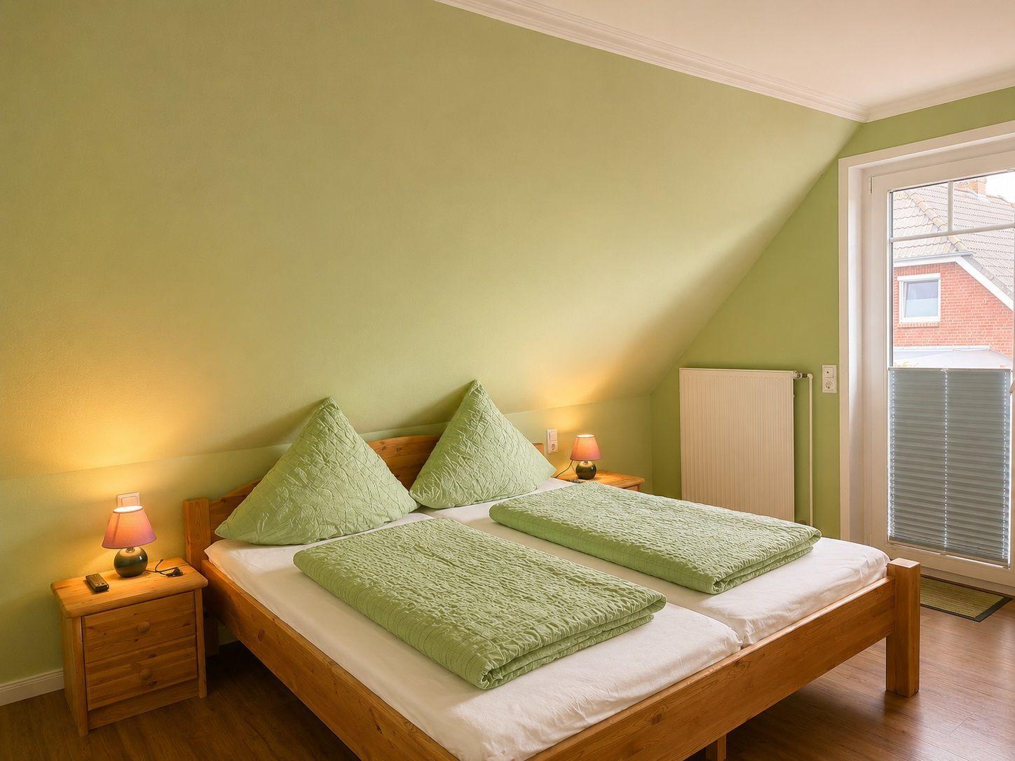 Schlafzimmer mit Doppelbett, grünen Kissen und Decken, Holzgestell und zwei Nachttischen mit Lampen.