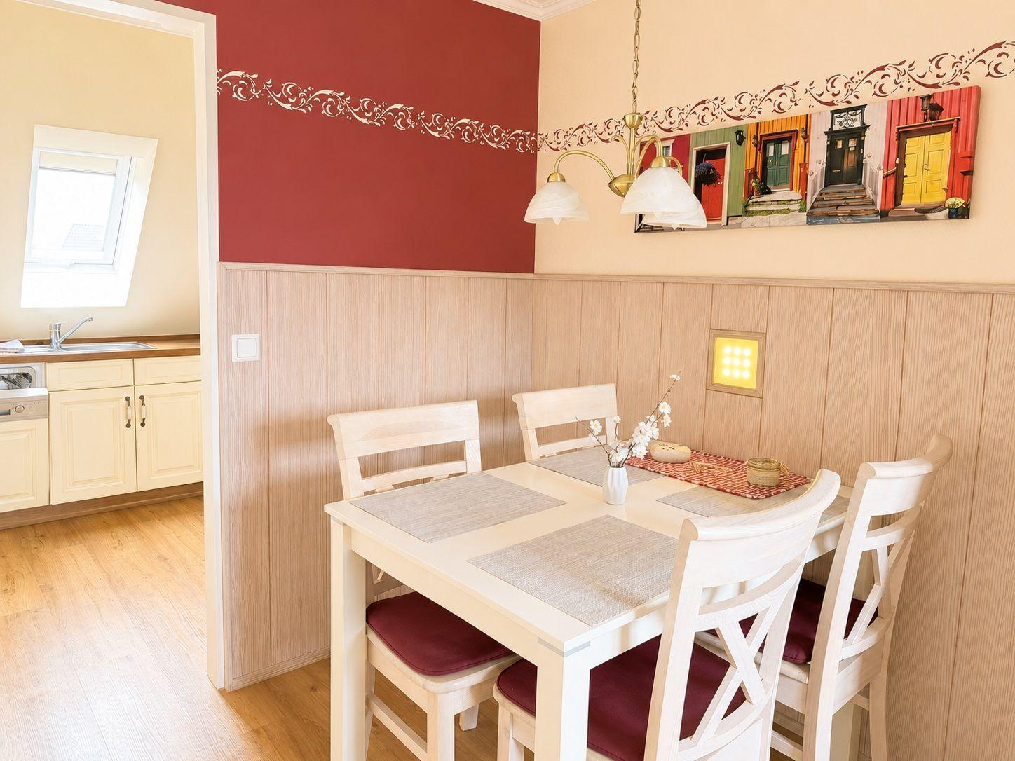 Esszimmer mit Tisch, Stühlen und Wanddekoration. Küche ist im Hintergrund sichtbar.