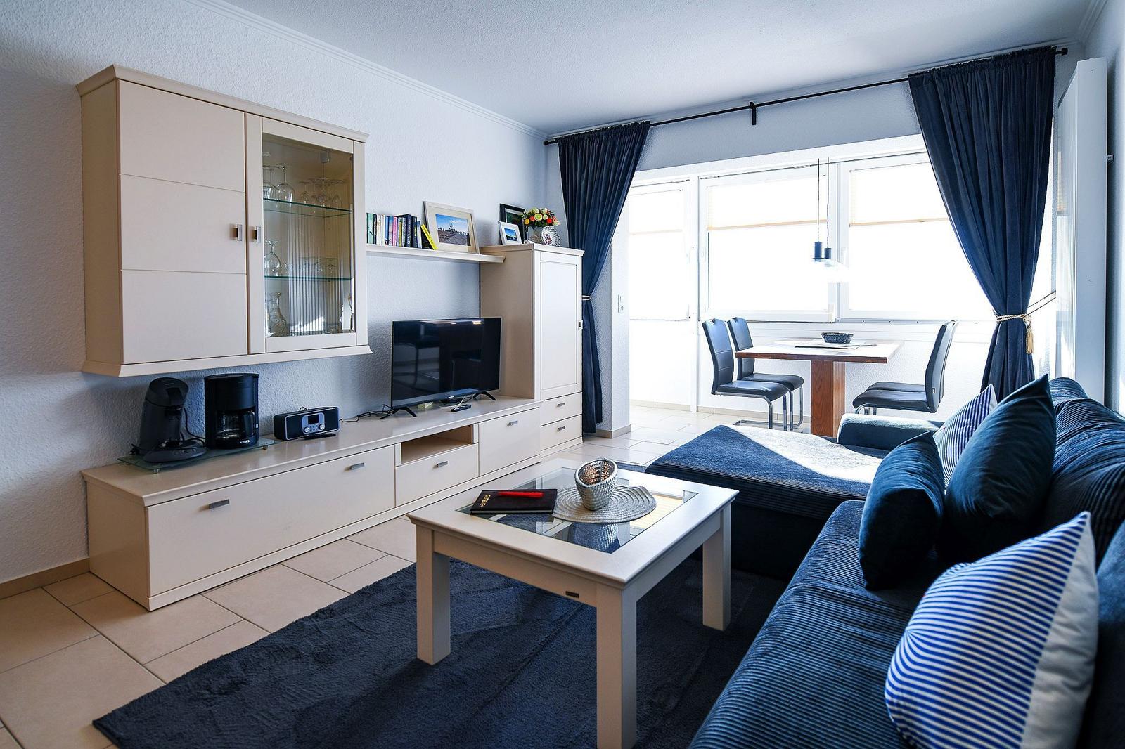 Wohnzimmer mit Sofa, Couchtisch und TV-Möbel. Fenster mit Vorhängen und Esstisch im Hintergrund.