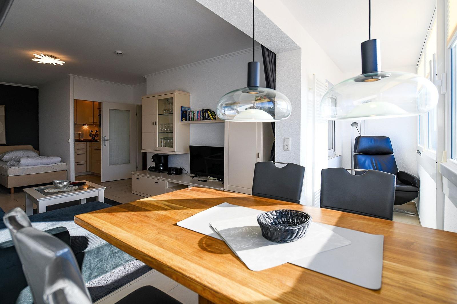 Esszimmer mit Holztisch, Stühlen und zwei Hängelampen. Im Hintergrund Küche und Schlafzimmer.
