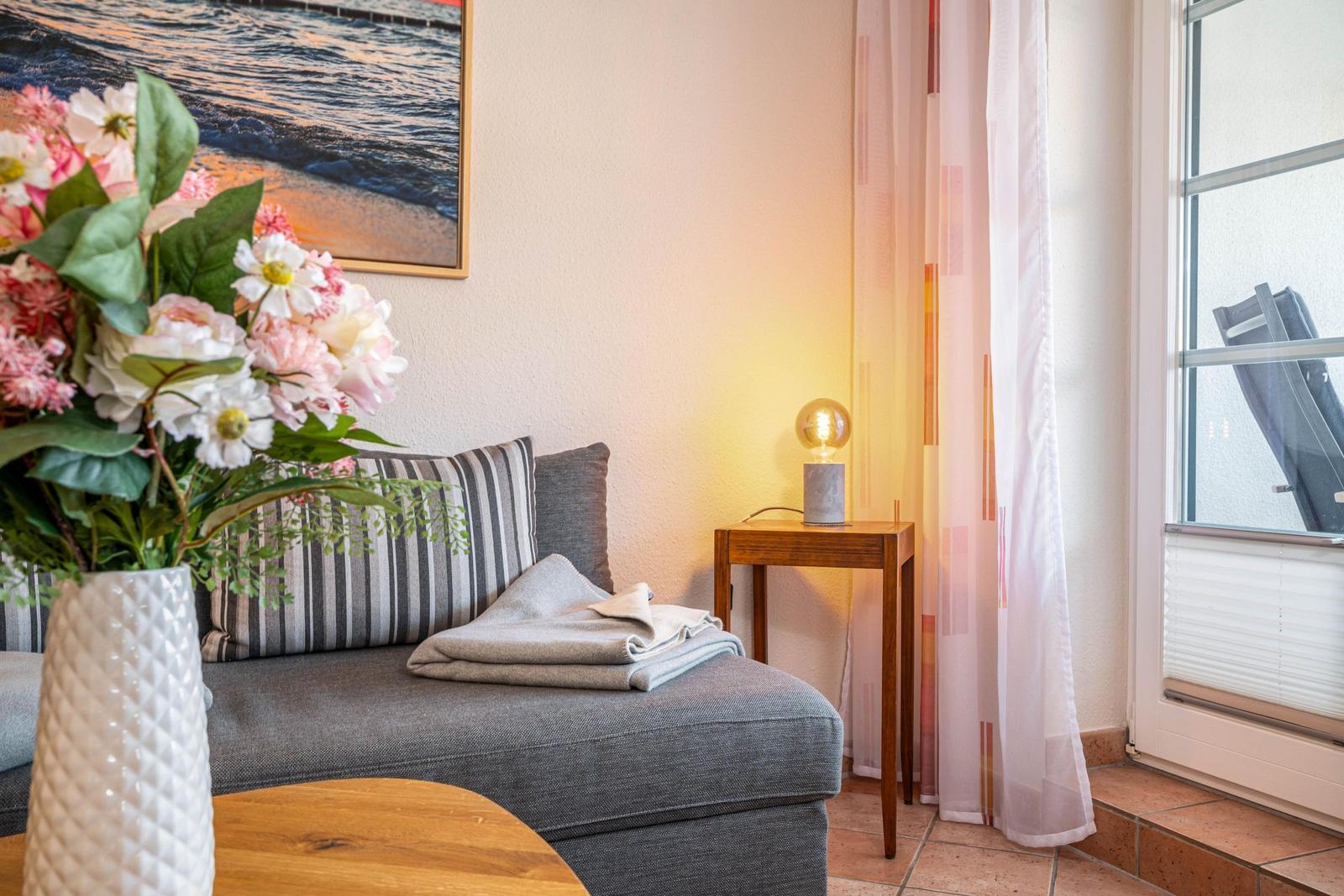 Graue Couch mit Kissen und Decke, daneben Tisch mit Lampe und Blumen.