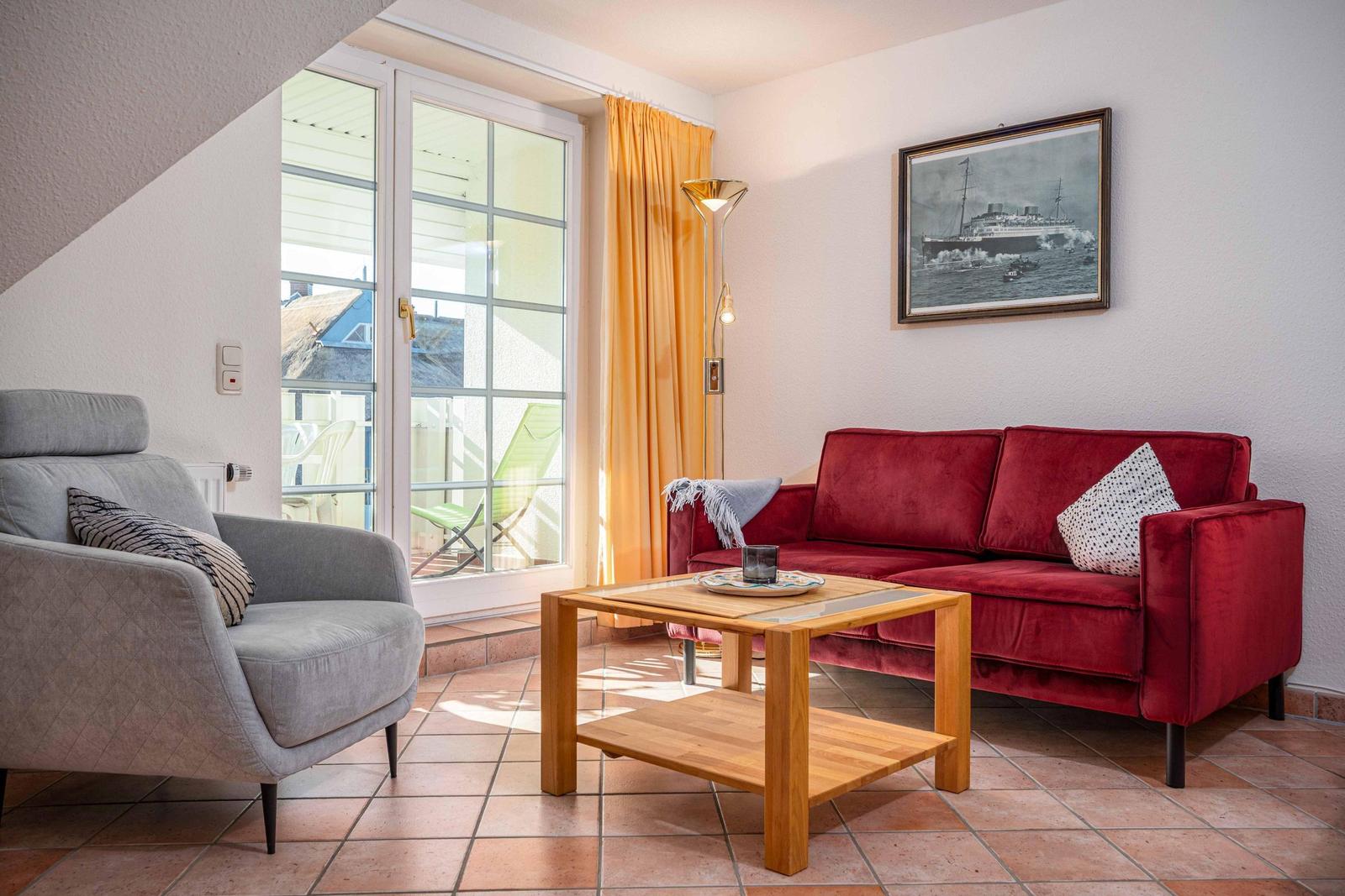 Ein Wohnzimmer mit rotem Sofa, grauem Sessel und Holztisch. Durchgehendes Fenster mit orangefarbenen Vorhängen.