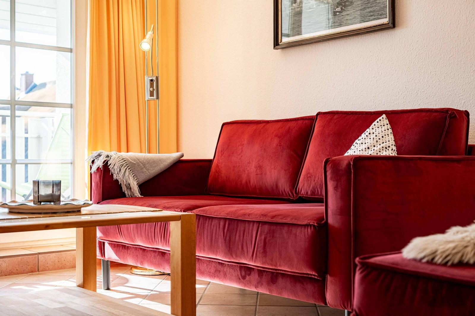 Roter Samtsofa mit Kissen und Decke vor Fenster mit orangefarbenen Vorhängen.