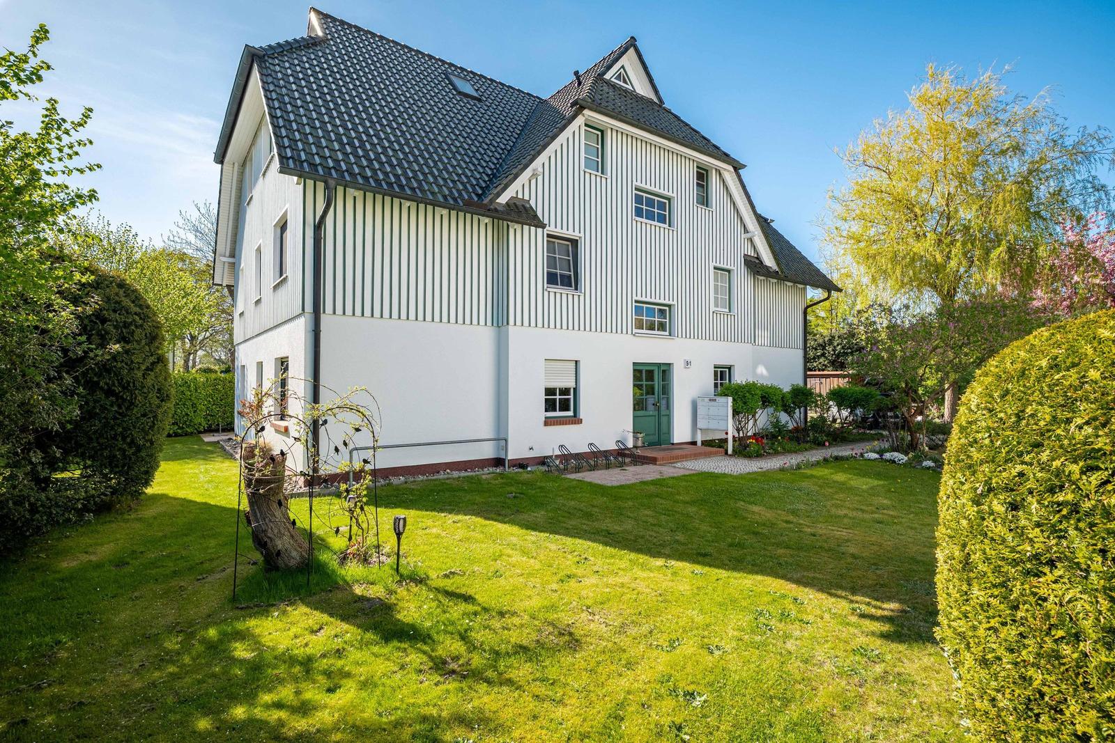 Haus mit weißer Fassade, grüner Tür und gepflegtem Garten unter blauem Himmel.