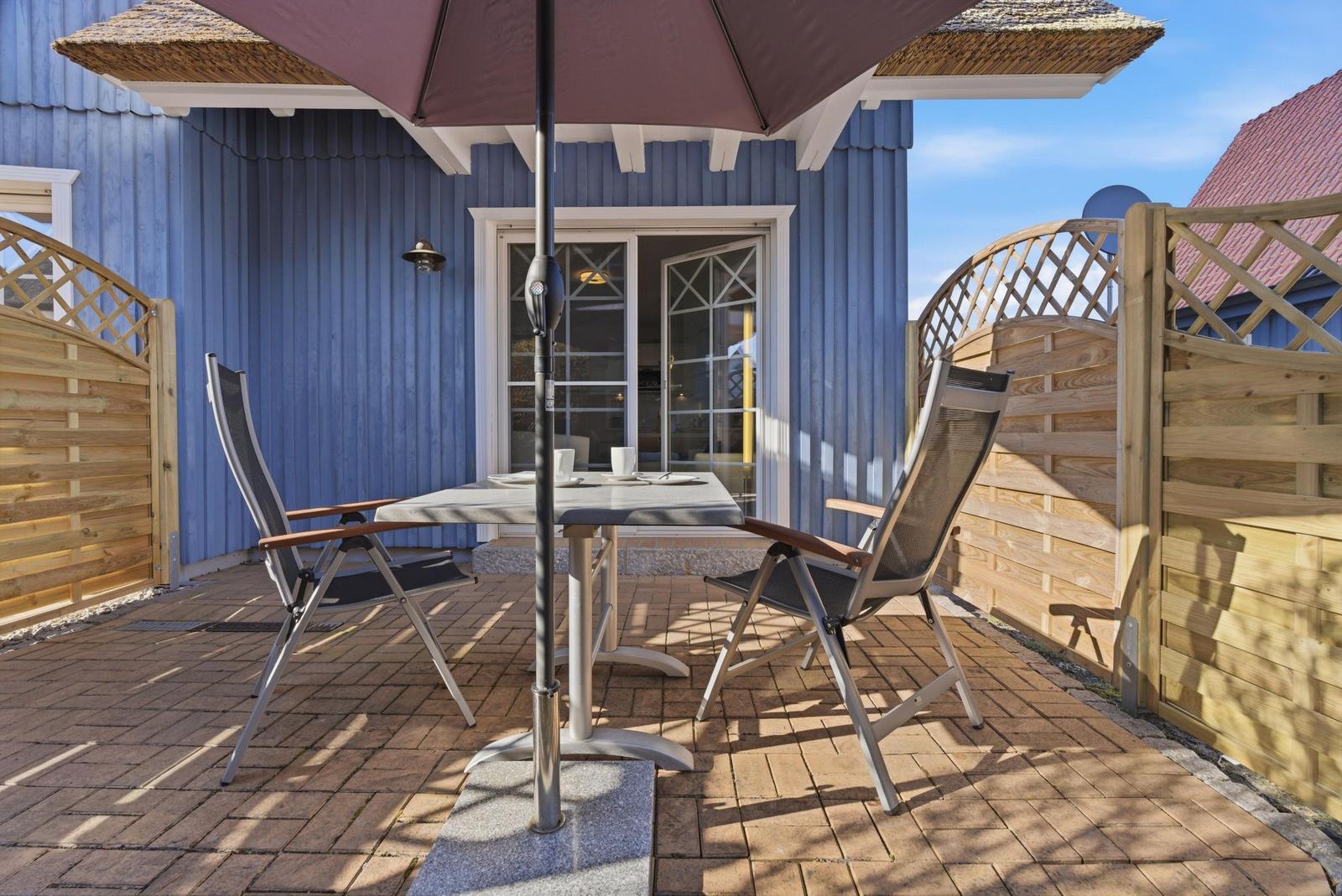 Terrasse mit Tisch, zwei Stühlen und Sonnenschirm vor blauem Holzhaus.