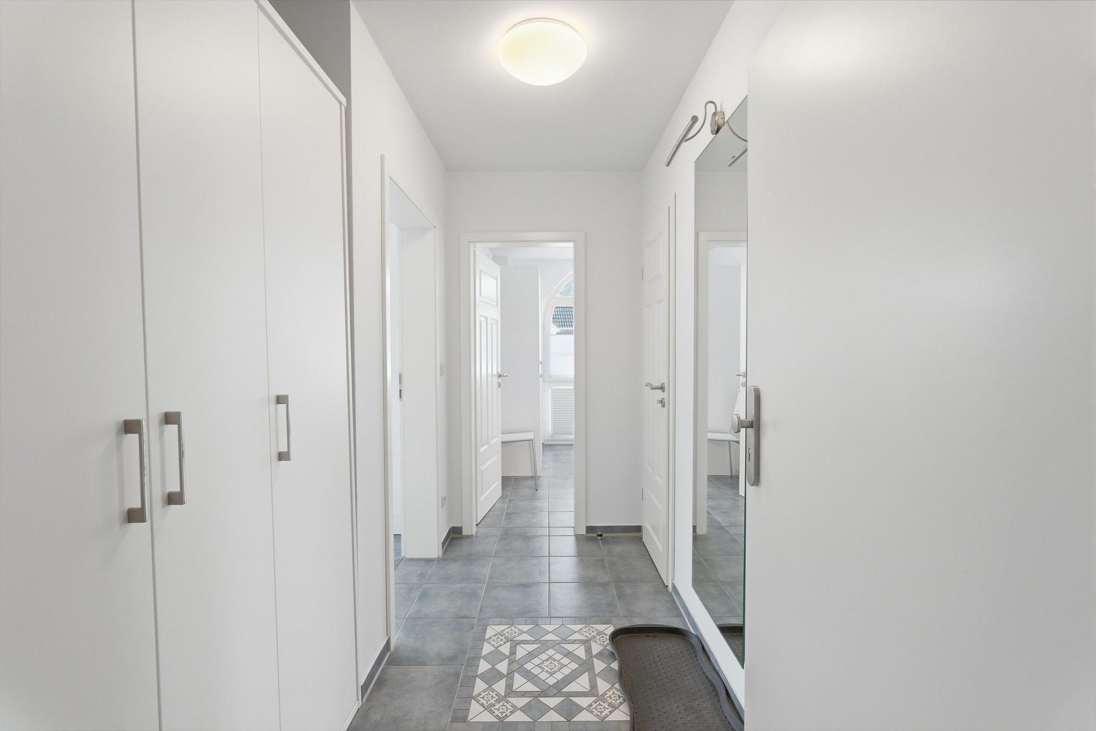 Flur mit weißen Wänden, grauem Fliesenboden und Spiegel. Links Garderobe, rechts Tür.