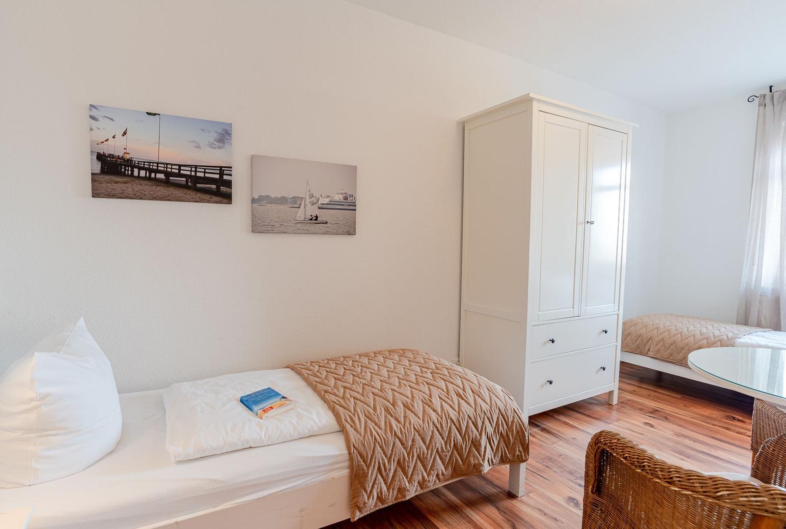 Zimmer mit zwei Betten, weißen Schränken und Holzboden. Wandbilder zeigen Hafen und Segelboot.