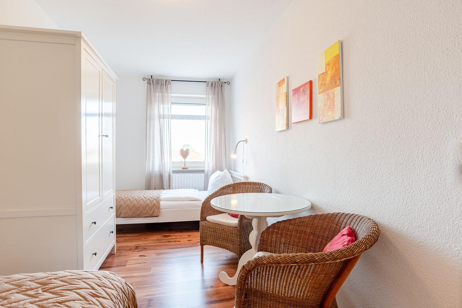Zimmer mit Bett, Schrank, Tisch und zwei Stühlen. Fenster mit Vorhängen und Wandbilder.