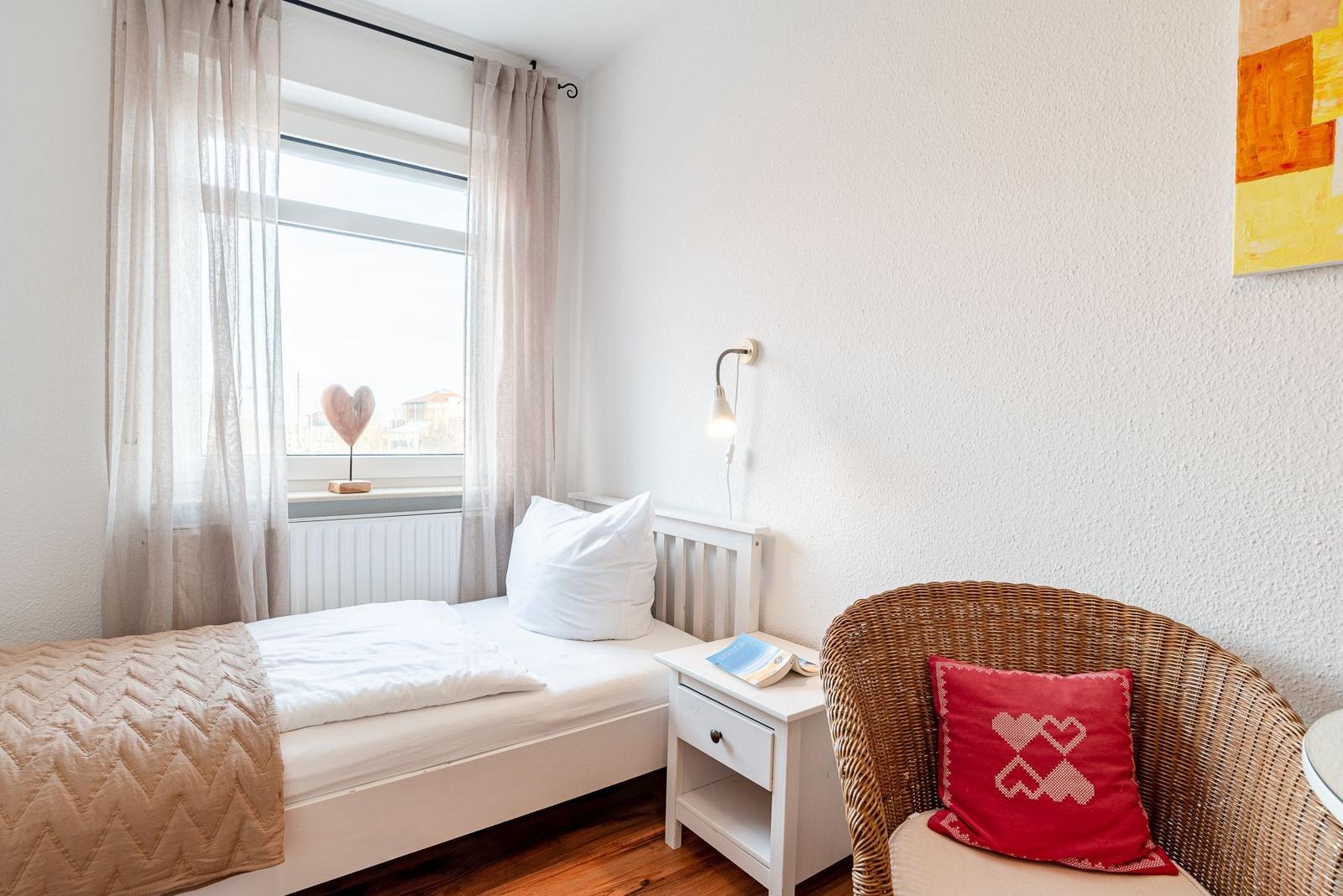 Ein Schlafzimmer mit Bett, Nachttisch und Stuhl. Fenster mit Vorhängen und Wandbild.