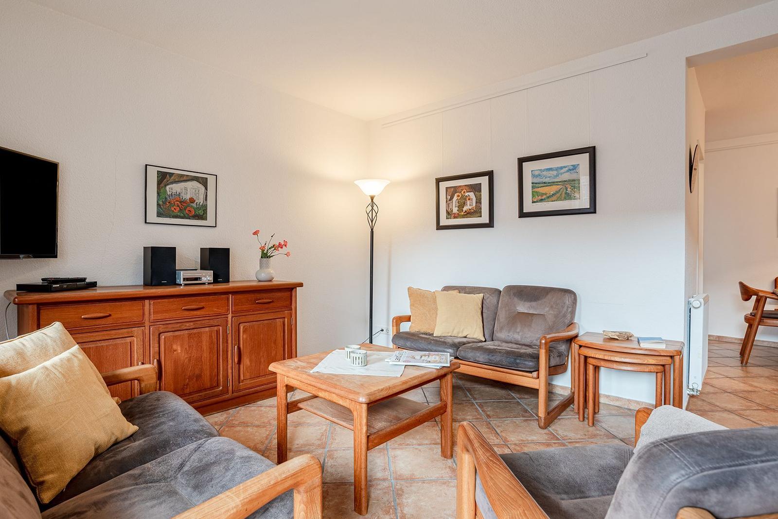 Wohnzimmer mit Holzmöbeln, Sofa, Couchtisch und Fernseher. Wandbilder und Stehlampe sichtbar.