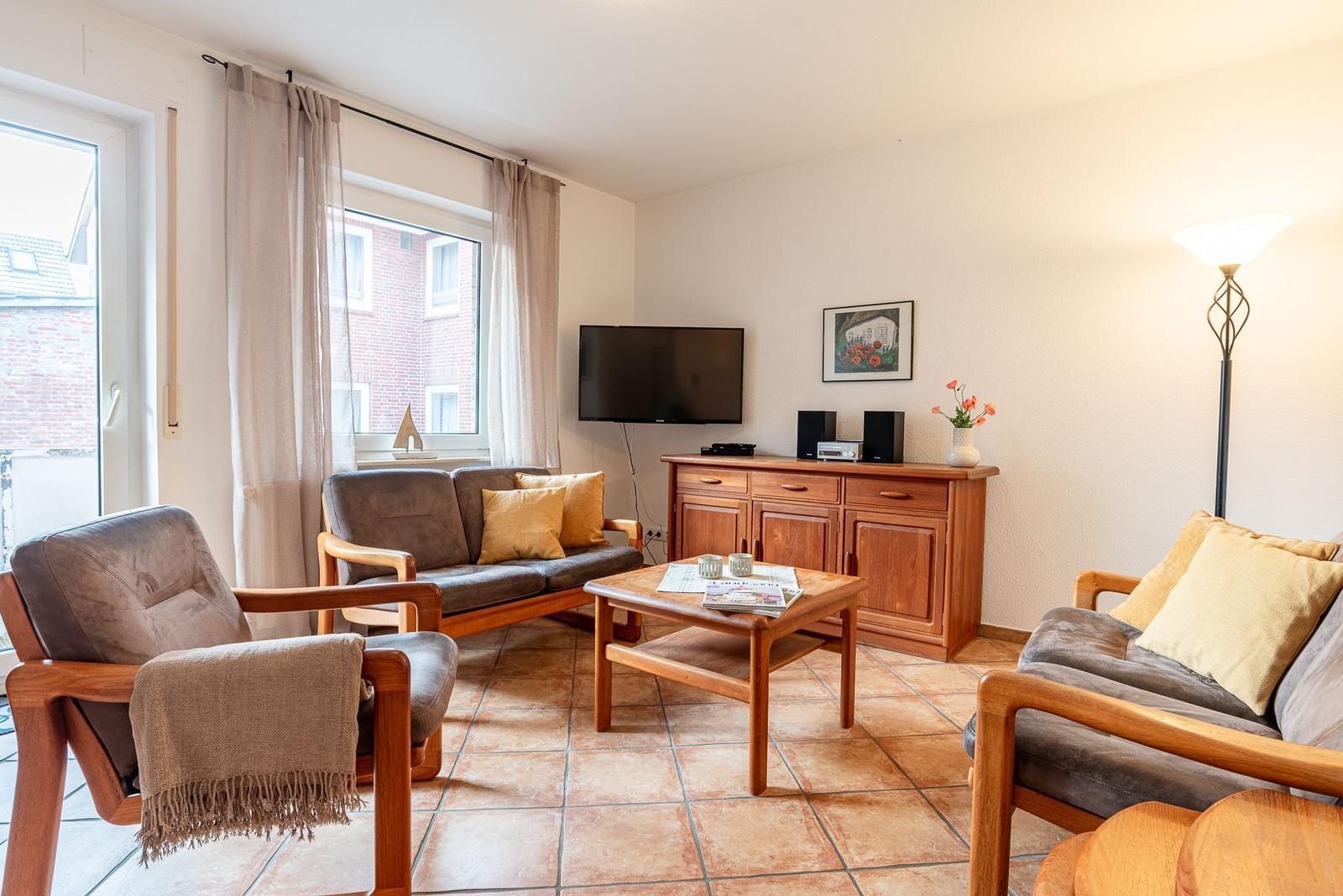 Wohnzimmer mit Sofa, Sessel, Couchtisch und Fernseher. Holzmöbel und Fenster mit Vorhängen.