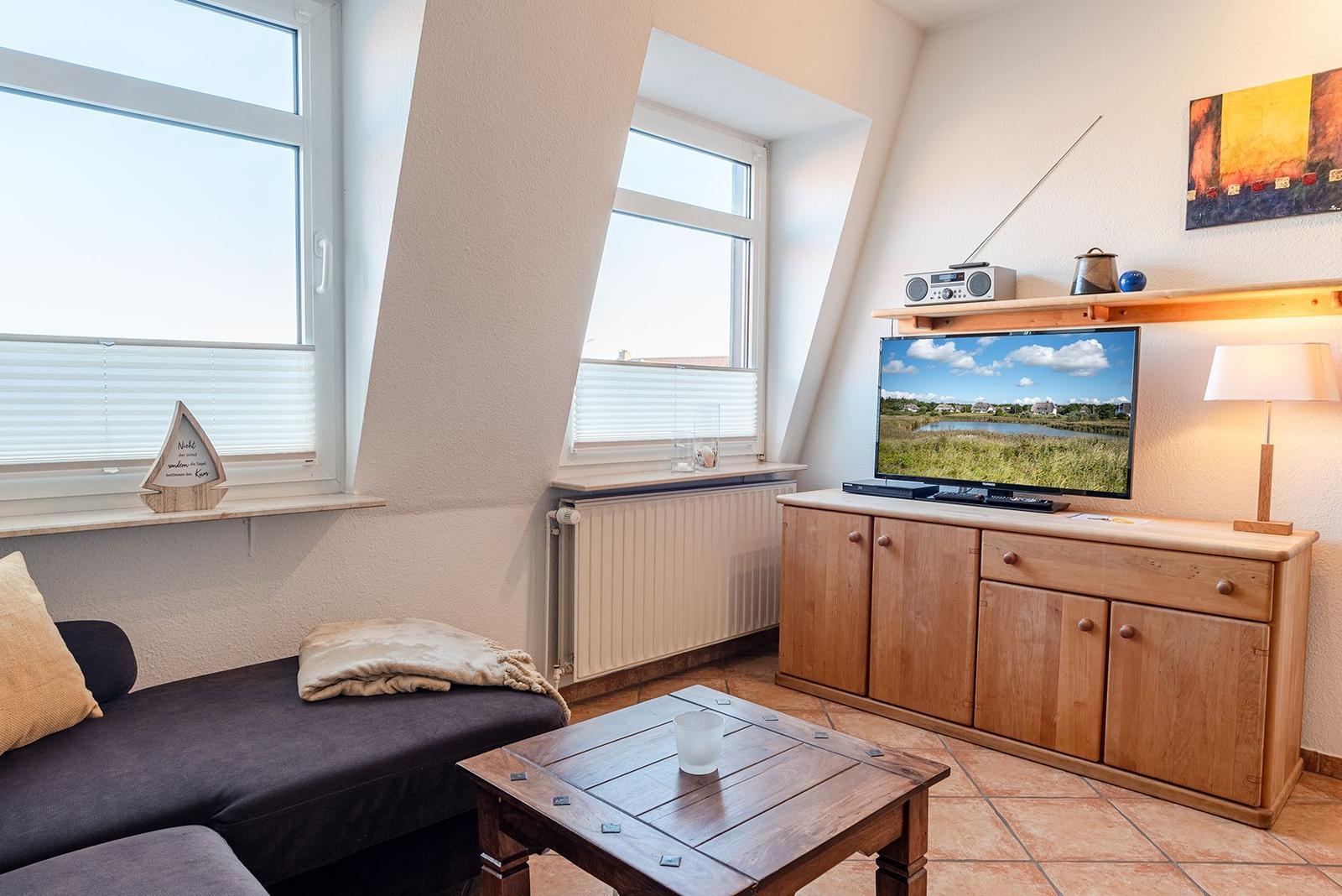Wohnzimmer mit TV, Holzmöbeln und Fenstern mit Jalousien.