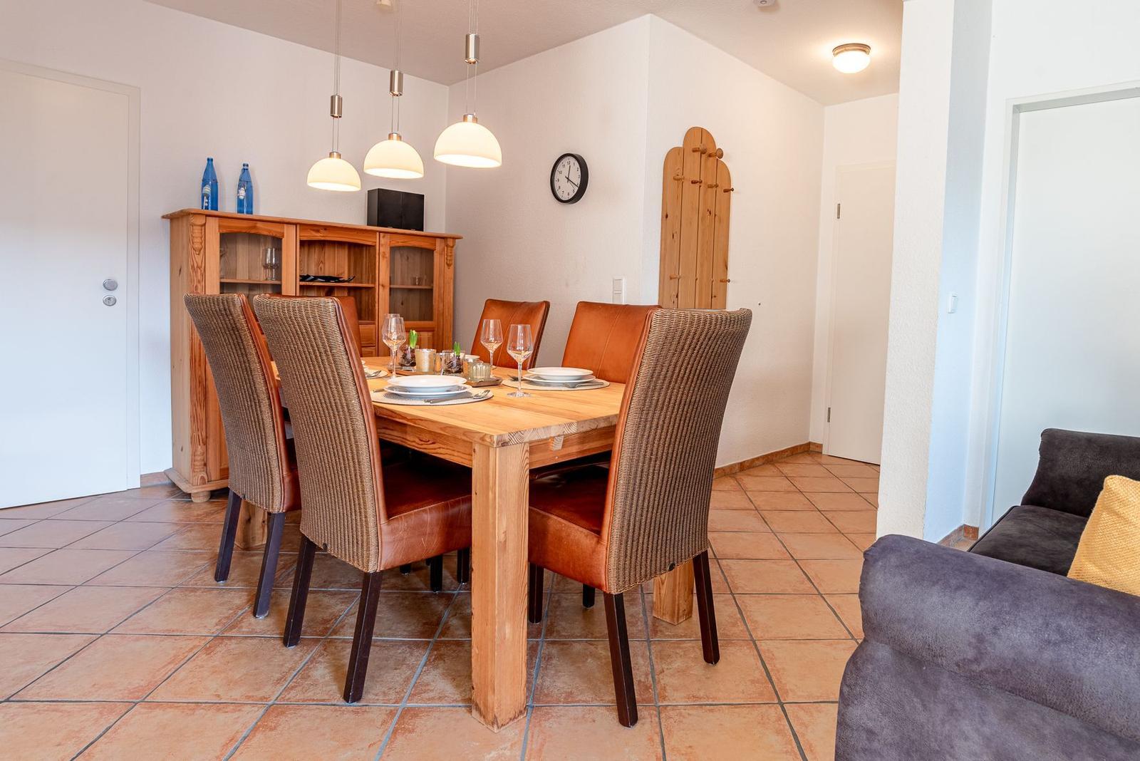 Essbereich mit Holztisch, Stühlen und Sideboard. Fliesenboden und Wanduhr sichtbar.