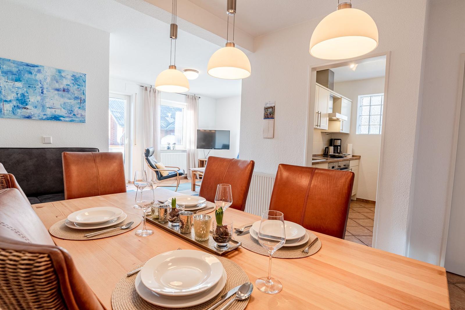 Esszimmer mit Holztisch, braunen Ledersitzen und hängenden Lampen. Küche und Wohnbereich sind sichtbar.