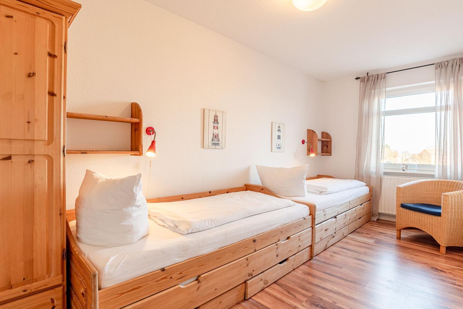 Zwei Holzbetten mit weißen Laken, ein Schrank und ein Sessel vor einem Fenster.