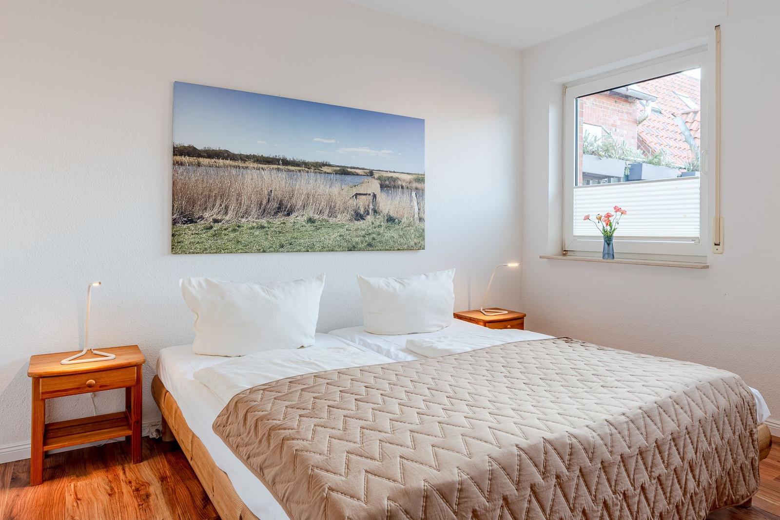 Schlafzimmer mit Doppelbett, Nachttisch und Wandbild. Fenster mit Blumen.