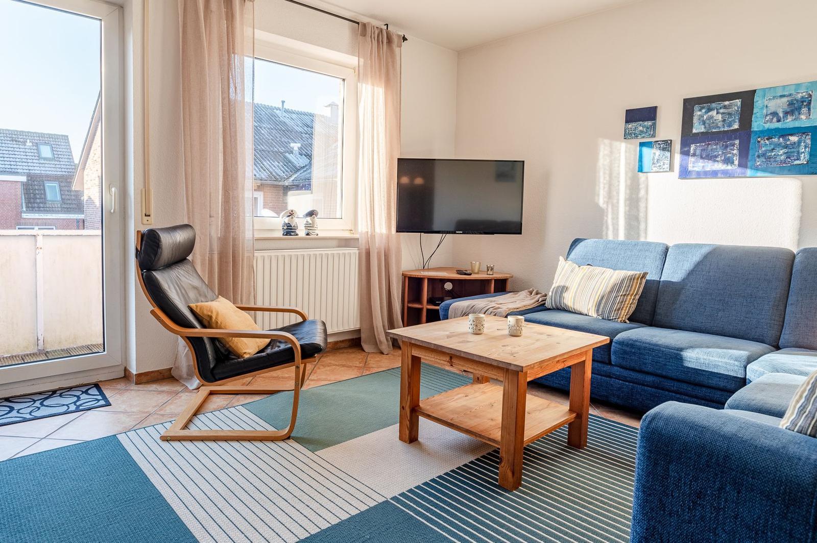 Ein Wohnzimmer mit blauem Sofa, Holztisch und Fernseher. Ein Fenster und eine Glastür führen auf eine Terrasse.