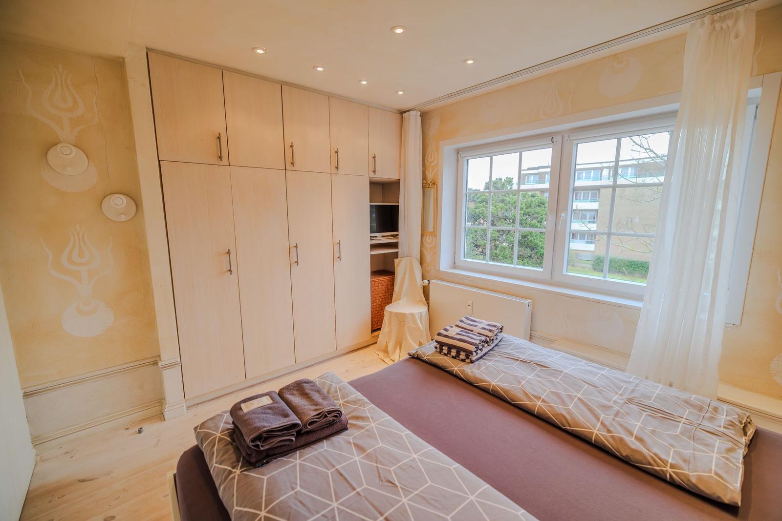 Schlafzimmer mit Bett, Kleiderschrank und Fenster. Wand mit Muster. Handtücher auf Bett.
