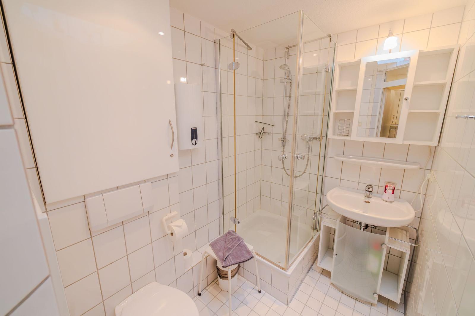 Badezimmer mit Dusche, Waschbecken und Toilette. Weiße Fliesen und Schrank.