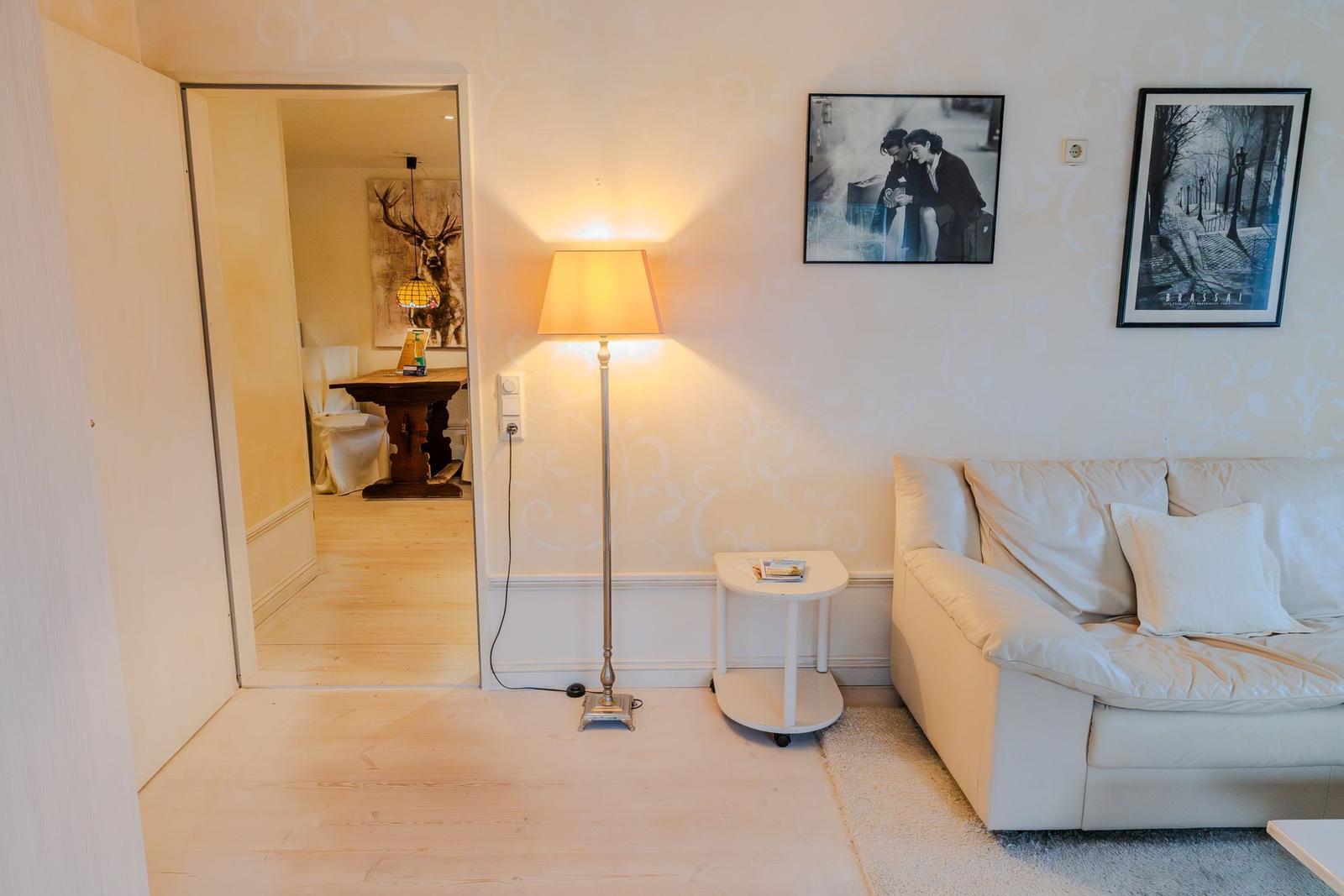 Wohnzimmer mit Sofa, Stehlampe und Bildern an der Wand. Durchgang zur Küche mit Tisch und Wandbild.