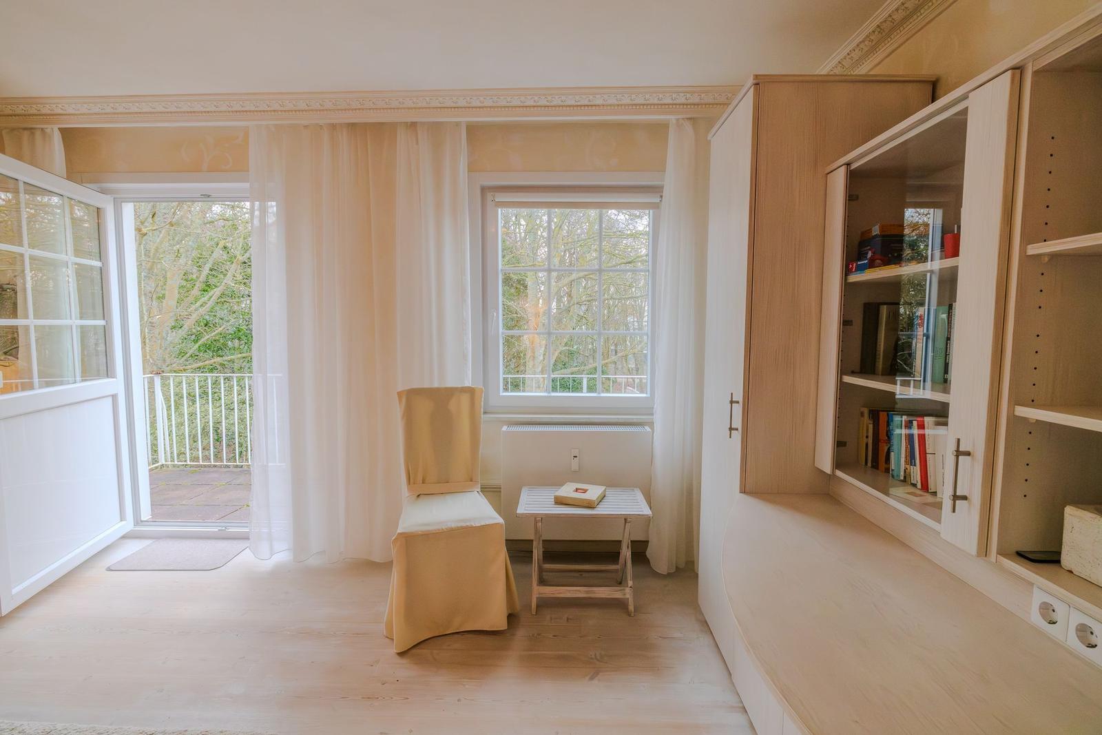 Ein Raum mit einem Fenster, einer Tür zum Balkon und einem Bücherregal.