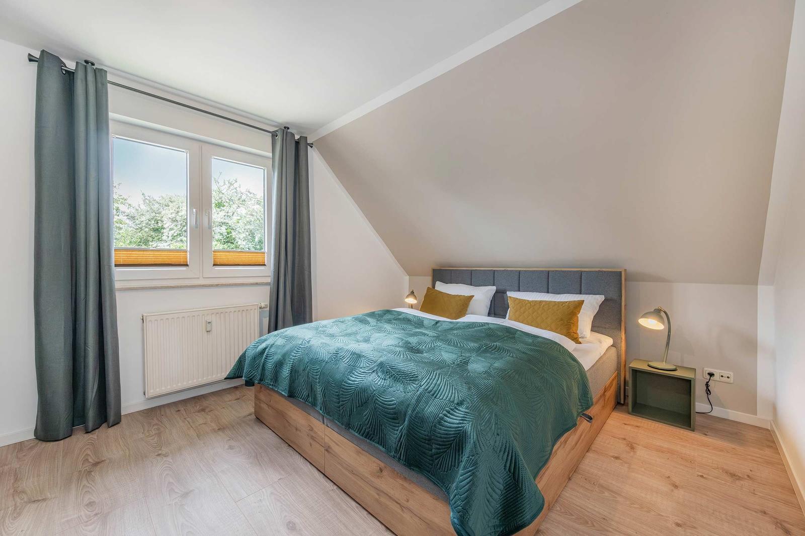Schlafzimmer mit Dachschrägen, Bett mit grünem Überwurf und Fenster mit Blick auf Bäume.