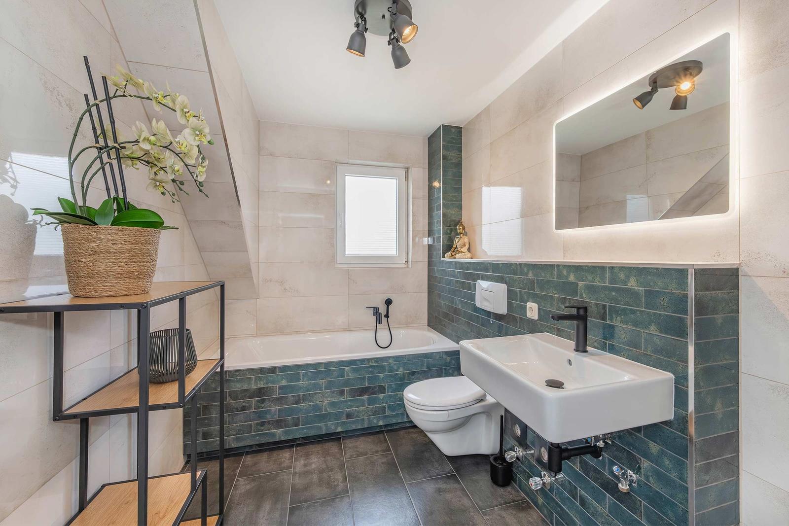 Bad mit Badewanne, Waschbecken und Toilette. Wandverkleidung in dunkelblau und hellbeige Fliesen.