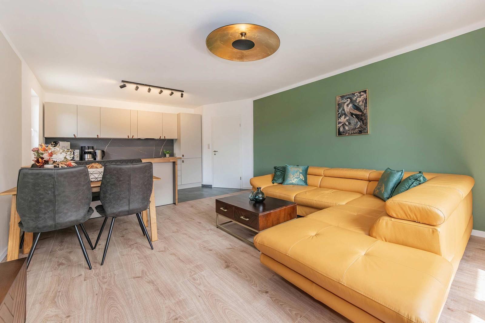 Wohnzimmer mit gelbem Sofa, grüner Wand und Essbereich mit Holzboden.
