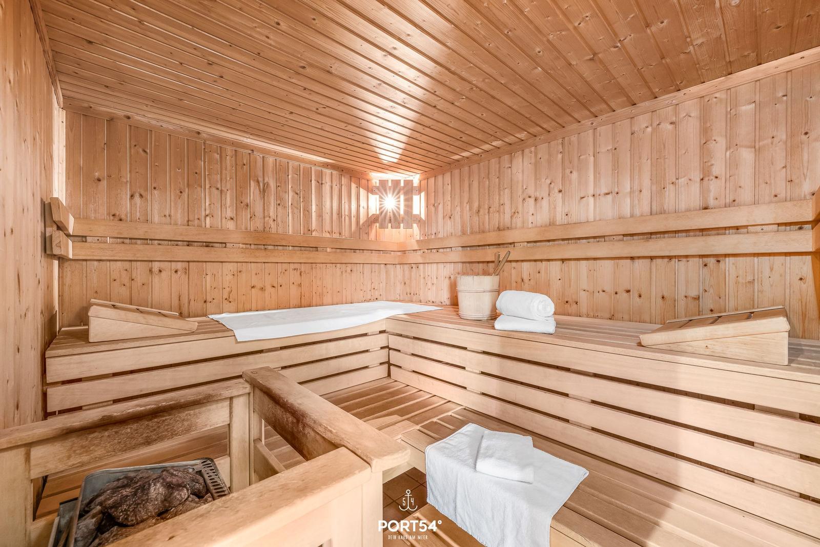 Sauna mit Holzbank, Handtuch und Eimer. Licht fällt durch Fenster.