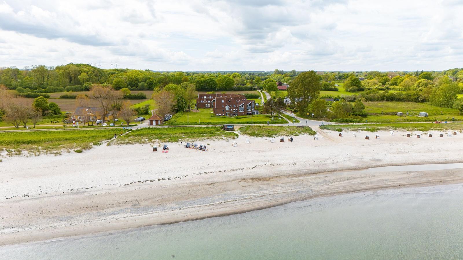 Strand mit Dünen, Häusern und Grünflächen im Hintergrund.