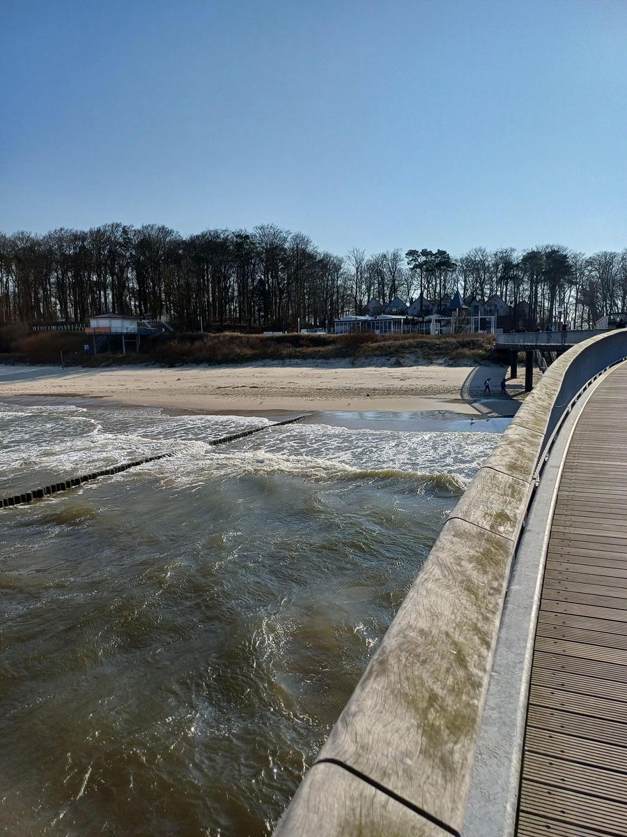 Holzbrücke führt zum Strand. Im Hintergrund sind Bäume und Gebäude zu sehen.